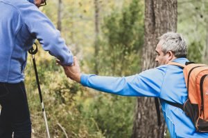 An older man helping another older man walk through a tricky forest