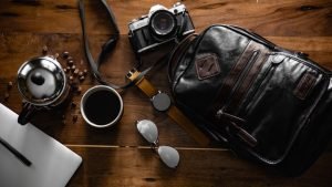 A backpack, a camera a watch and coffee on a table.